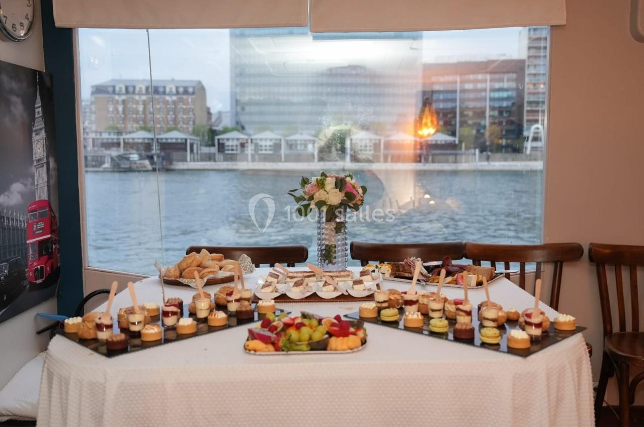 Table dressée avec des desserts, fruits, sandwichs et un bouquet de fleurs, devant une baie vitrée donnant sur une rivière.