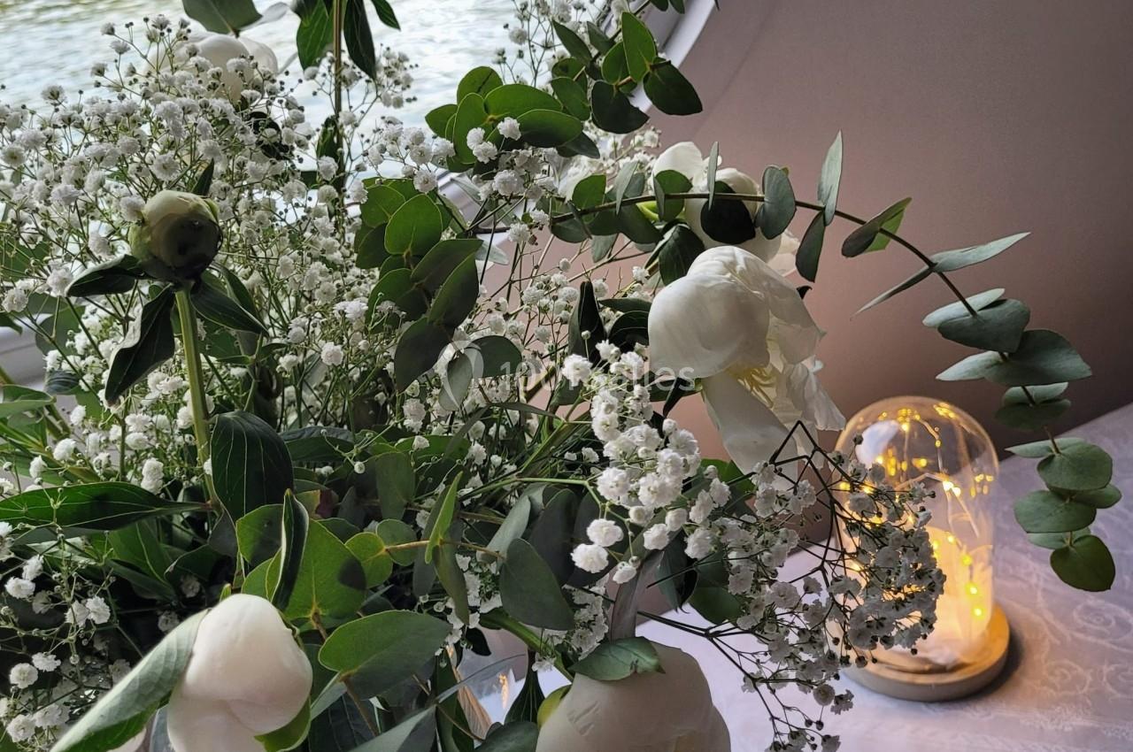 Bouquet de fleurs blanches et feuillage vert posé sur une table près d'une cloche lumineuse et d'une fenêtre.