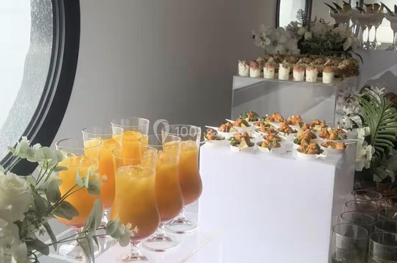 Buffet avec cocktails orange, verrines et bouchées salées, décoré de fleurs blanches et verdure.