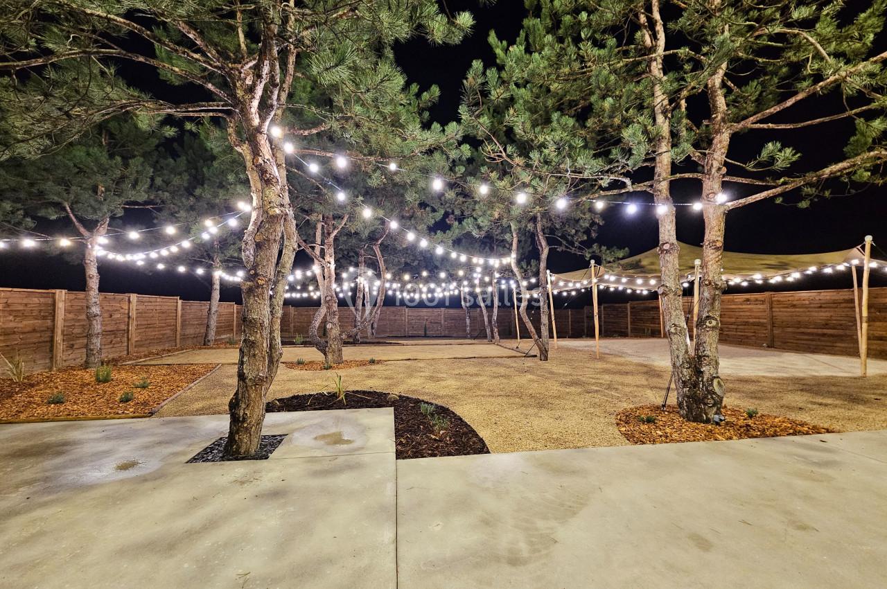 Espace extérieur de nuit avec des arbres éclairés par des guirlandes lumineuses suspendues, sol en béton et gravier.