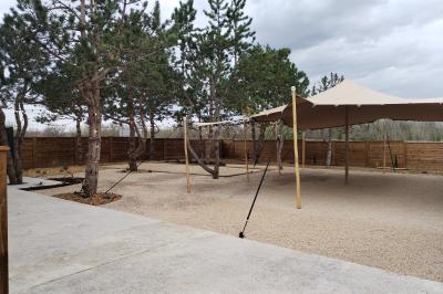 Espace extérieur avec sol en gravier, arbres, tente beige tendue et clôture en bois sous un ciel nuageux.