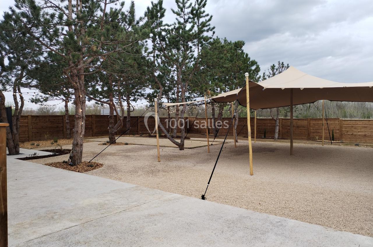 Espace extérieur avec sol en gravier, arbres, tente beige tendue et clôture en bois sous un ciel nuageux.