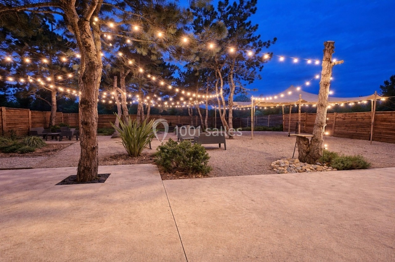 Cour extérieure avec guirlandes lumineuses suspendues entre des arbres, entourée de mobilier et de clôtures en bois.
