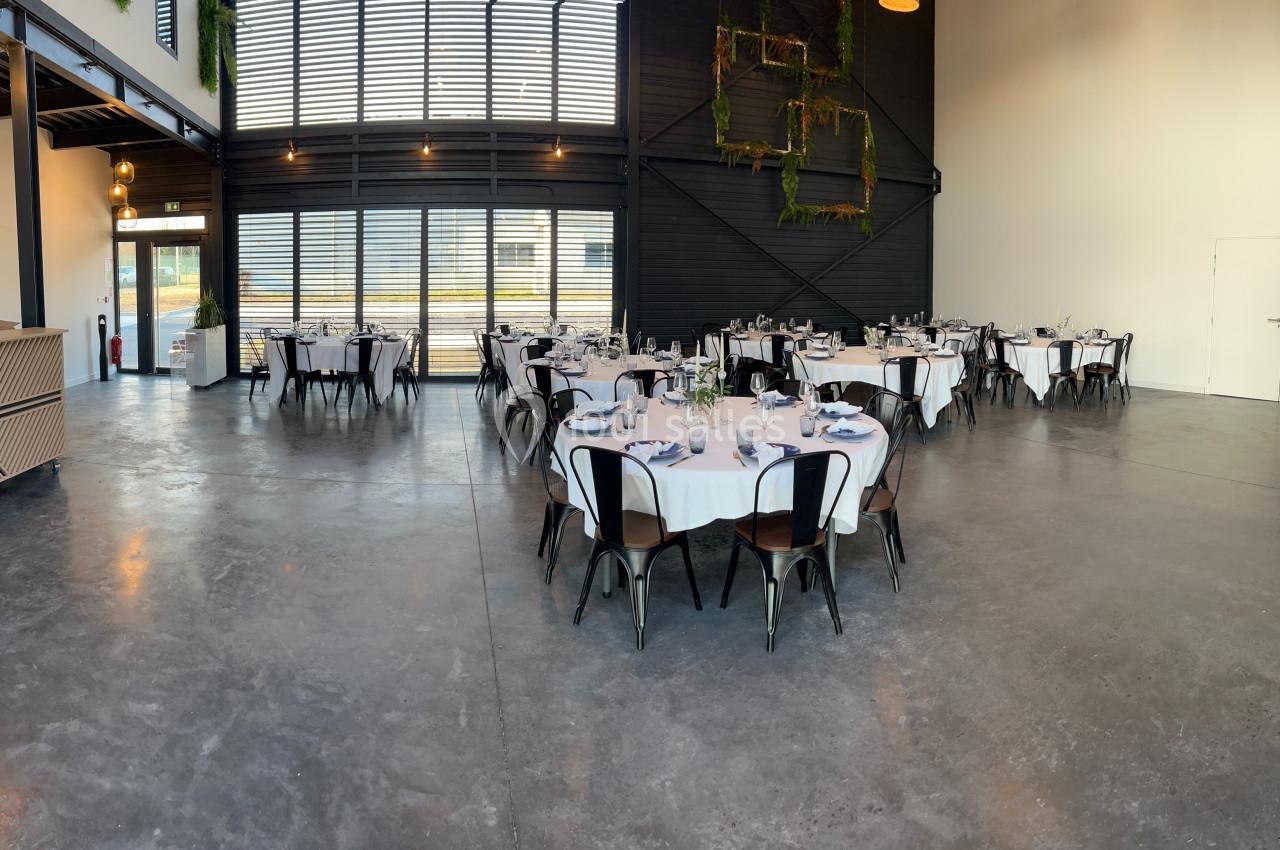 Salle de réception lumineuse avec tables rondes dressées, chaises noires et décorations végétales sur les murs.