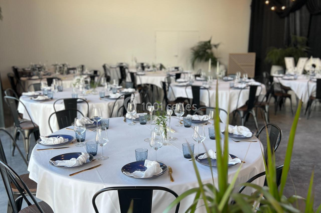 Salle de réception décorée avec des tables rondes dressées, nappes blanches, chaises noires et éclairage chaleureux.