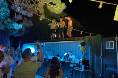 Espace extérieur avec sol en gravier, arbres, tente beige tendue et clôture en bois sous un ciel nuageux.