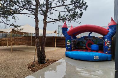 Espace extérieur avec sol en gravier, arbres, tente beige tendue et clôture en bois sous un ciel nuageux.