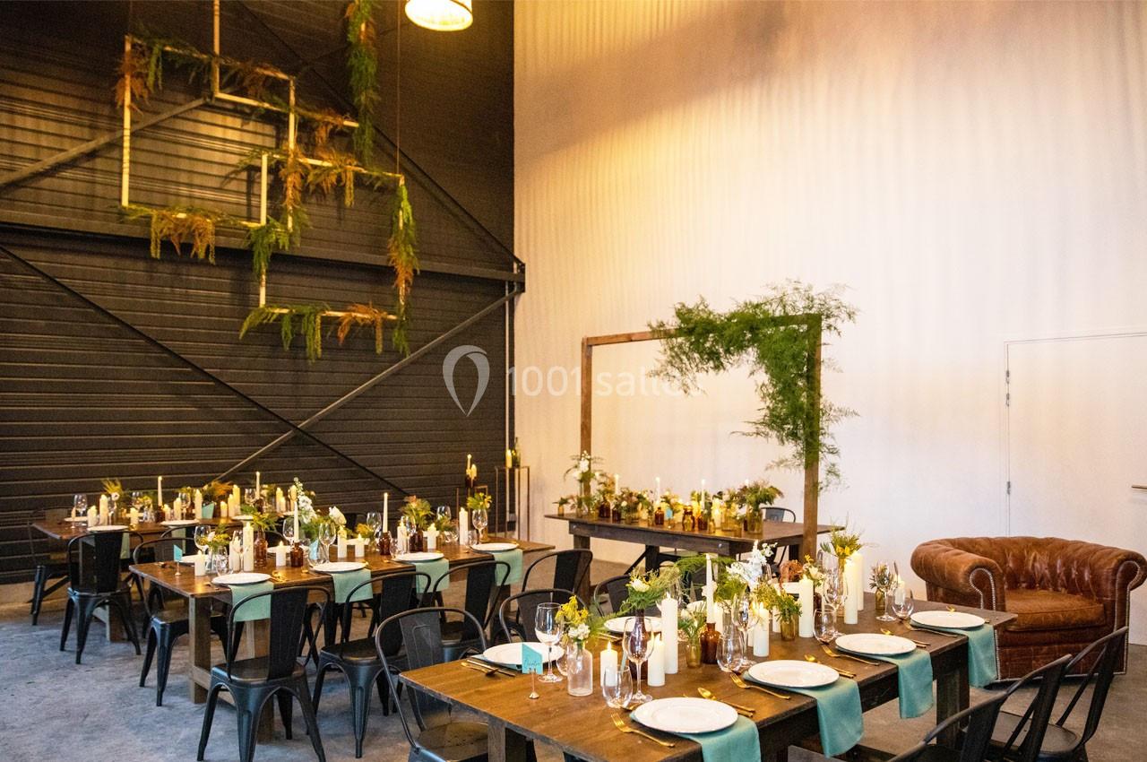 Salle décorée pour un événement, avec tables en bois, chaises noires, plantes suspendues et éclairage chaleureux.
