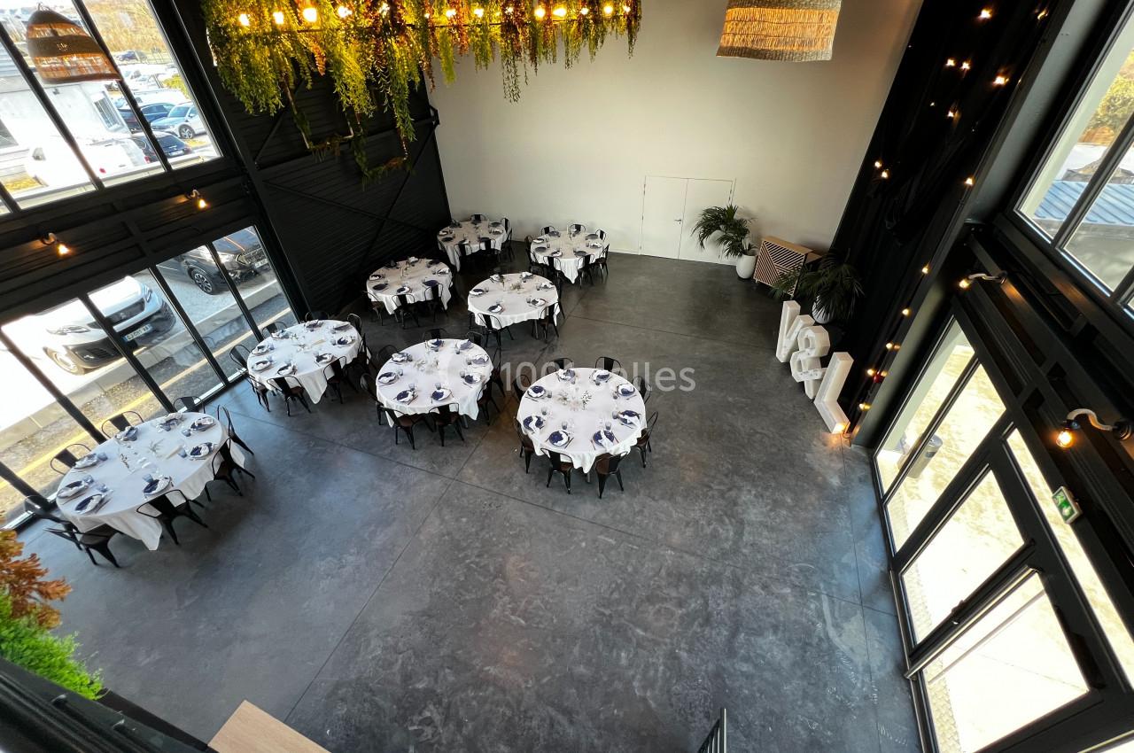 Salle de réception moderne avec tables rondes dressées, grandes baies vitrées et décoration végétale suspendue.