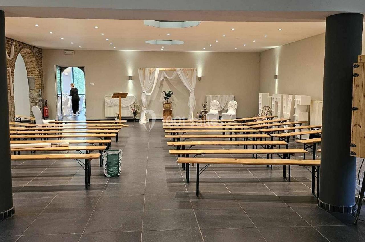 Salle de réception avec bancs en bois alignés, décorations blanches et colonnes noires, vue vers une entrée lumineuse.