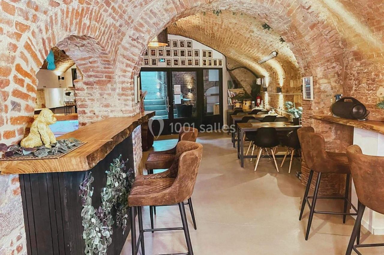 Salle voûtée en briques avec un bar en bois, des tables et des chaises, décorée de plantes et éclairée naturellement.