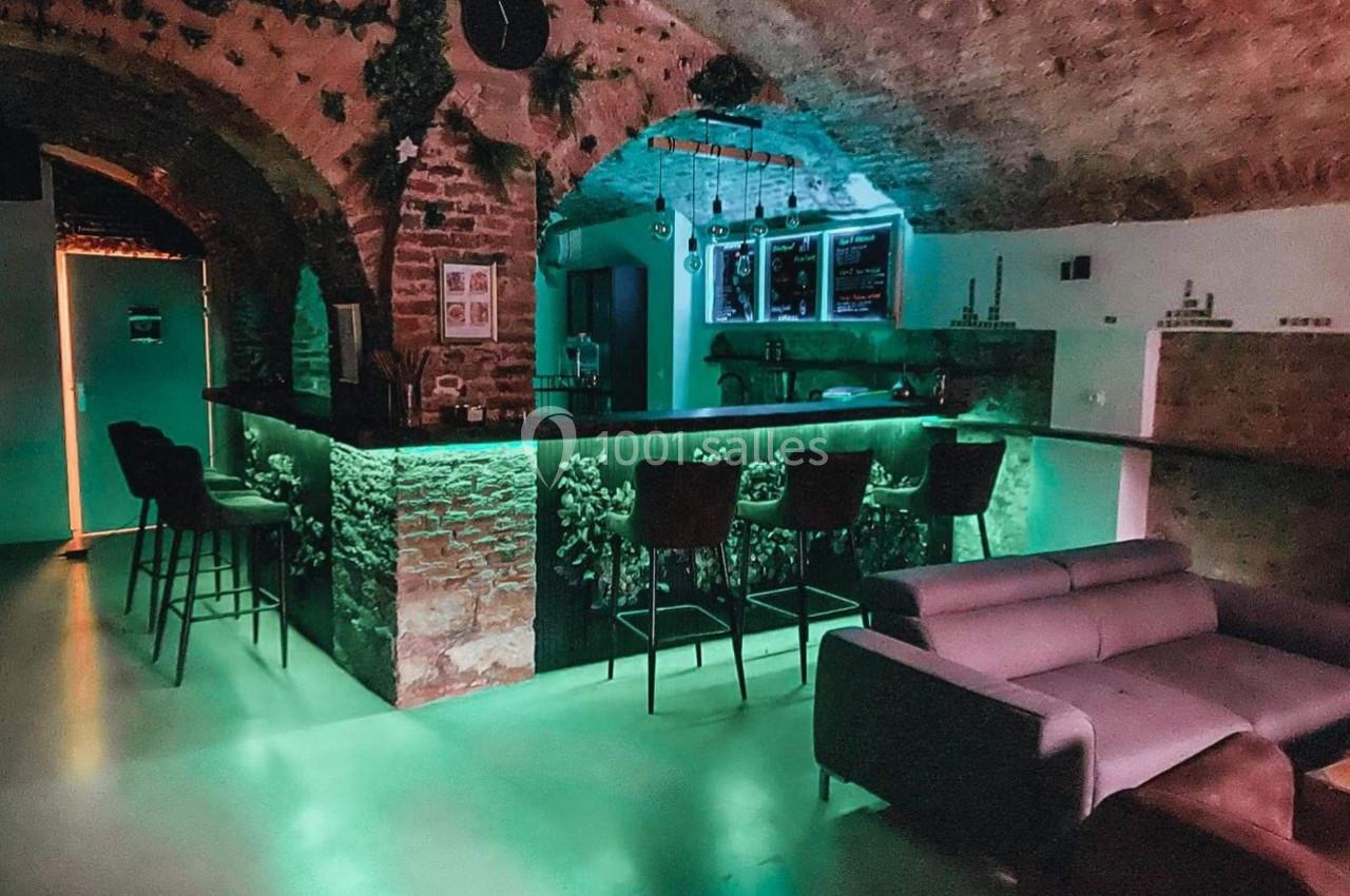 Bar avec comptoir en briques éclairé de lumières vertes, chaises hautes, canapé et voûtes en pierre apparente.