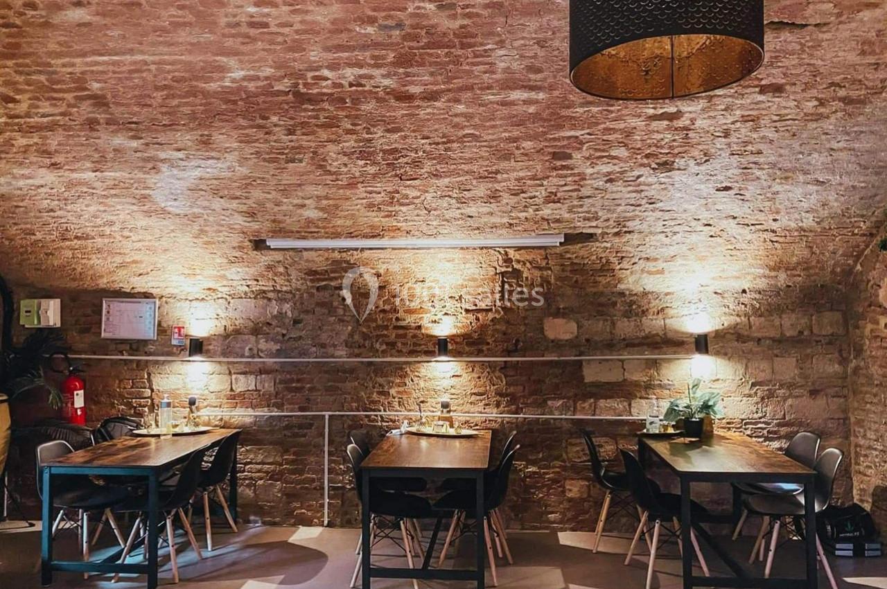 Salle voûtée en briques avec des tables en bois, des chaises noires et un éclairage tamisé.