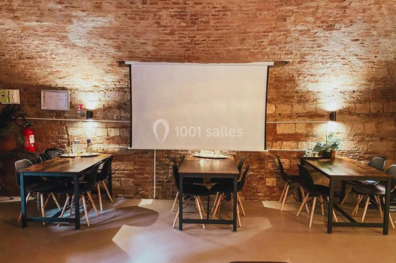 Salle voûtée en briques avec tables en bois, chaises noires et écran blanc au centre.