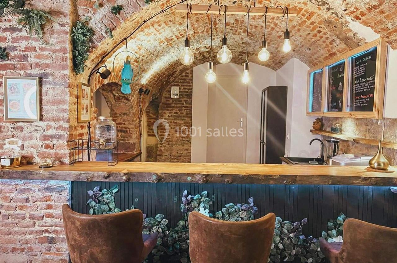 Bar avec comptoir en bois, tabourets rembourrés, murs en briques et plafond voûté éclairé par des ampoules suspendues.