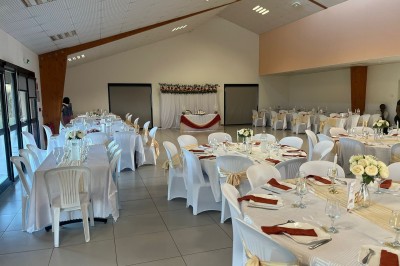Salle de réception décorée avec des tables dressées, nappes blanches, serviettes rouges et centre de table fleuri.