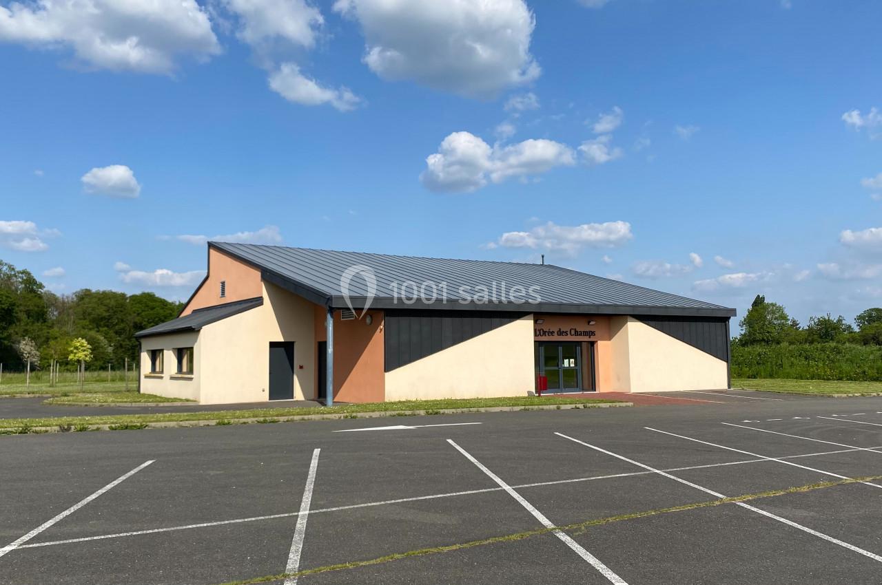 Bâtiment moderne avec toit en métal, entouré de verdure, vu depuis un parking vide sous un ciel bleu avec quelques nuages.