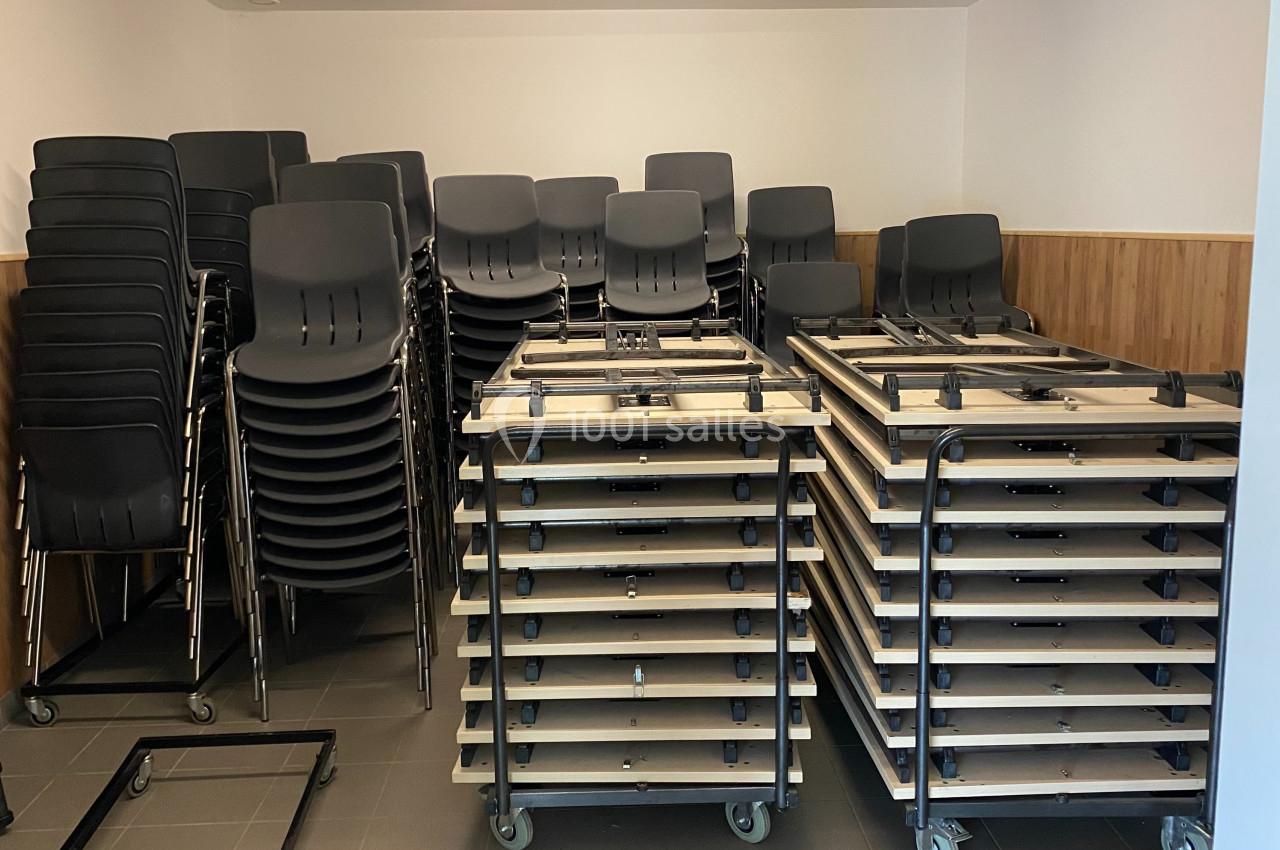 Chaises empilées et tables pliantes sur roulettes rangées dans une pièce aux murs clairs et au sol carrelé.