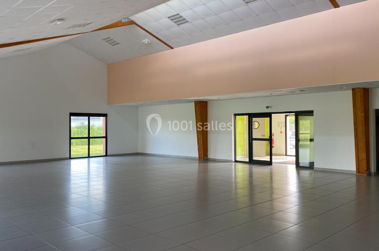 Salle vide avec carrelage gris, murs blancs, grandes fenêtres et portes vitrées laissant entrer la lumière naturelle.