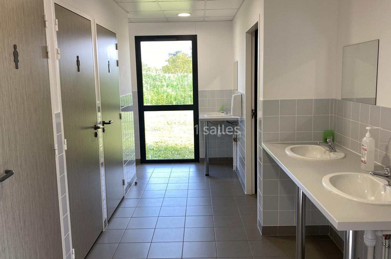 Vue d'un espace sanitaire avec lavabos, miroirs, cabines fermées et une fenêtre donnant sur un extérieur verdoyant.