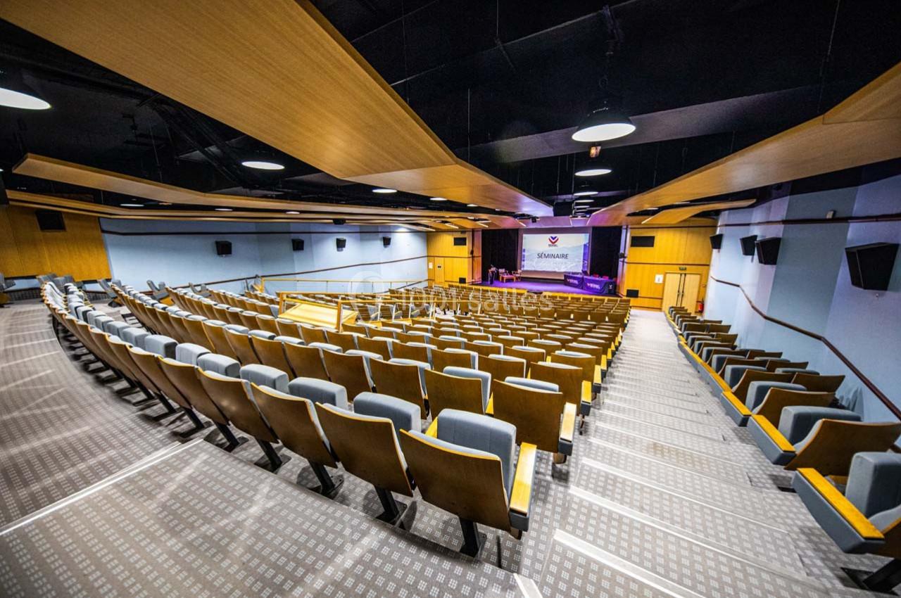 Salle de conférence moderne avec rangées de sièges rembourrés, scène équipée d'un écran et éclairage au plafond.