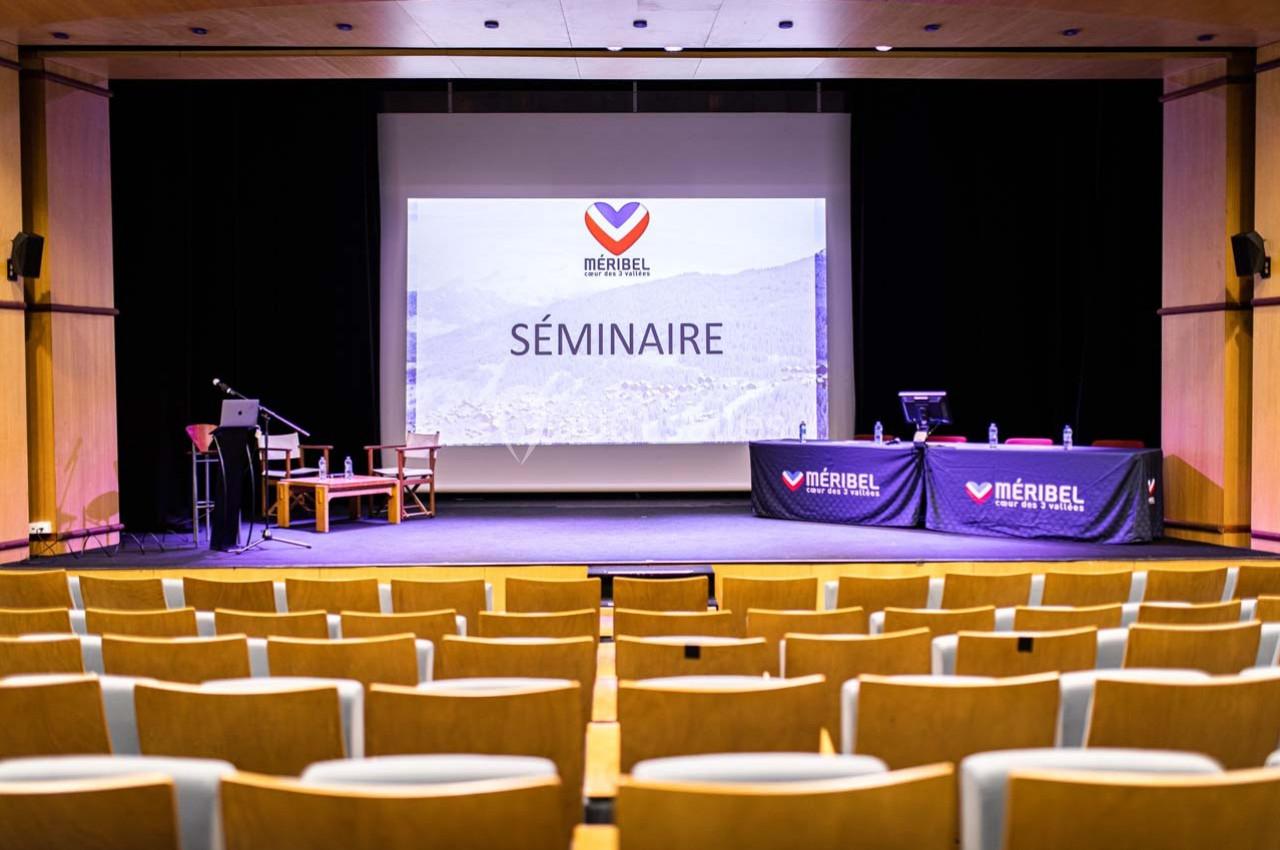 Salle de conférence avec écran de projection affichant ’Séminaire’, chaises alignées et table équipée sur scène.