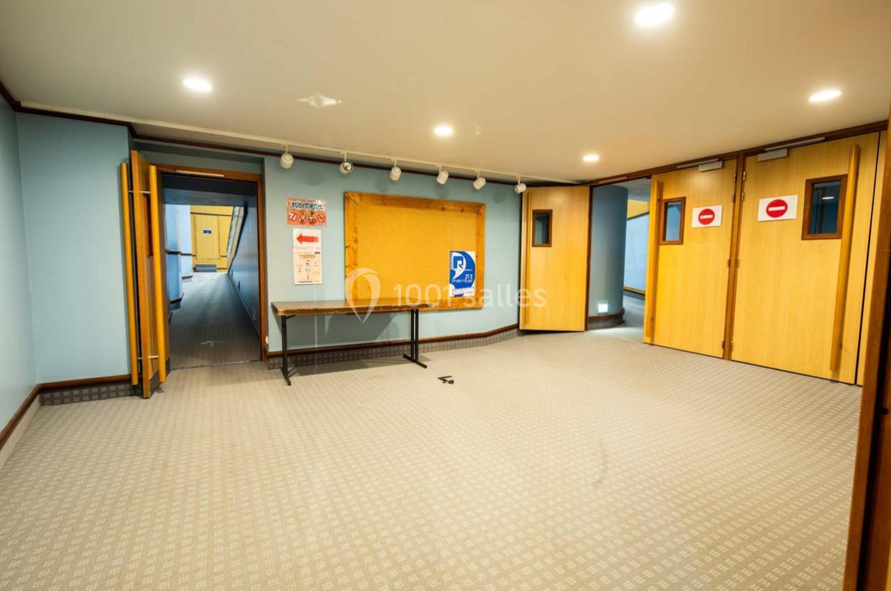 Salle vide avec murs bleu clair, sol en moquette beige, panneau d'affichage et portes en bois fermées.