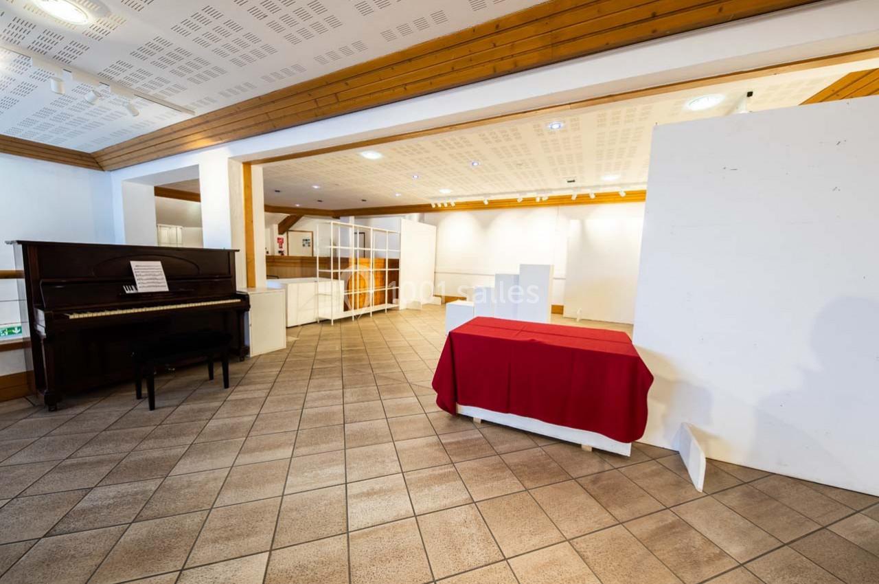 Salle vide avec carrelage au sol, murs blancs, un piano noir et une table recouverte d'une nappe rouge au premier plan.