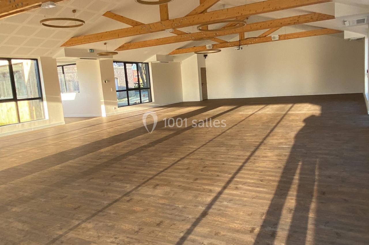 Salle lumineuse avec parquet en bois, poutres apparentes au plafond et grandes fenêtres laissant entrer la lumière naturelle.