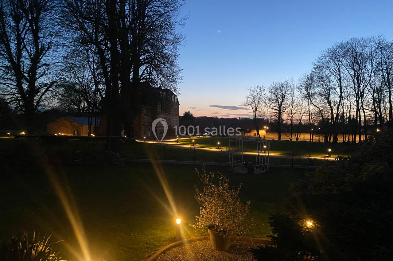 Jardin éclairé par des lampes au sol au crépuscule, avec des arbres et un bâtiment en arrière-plan.