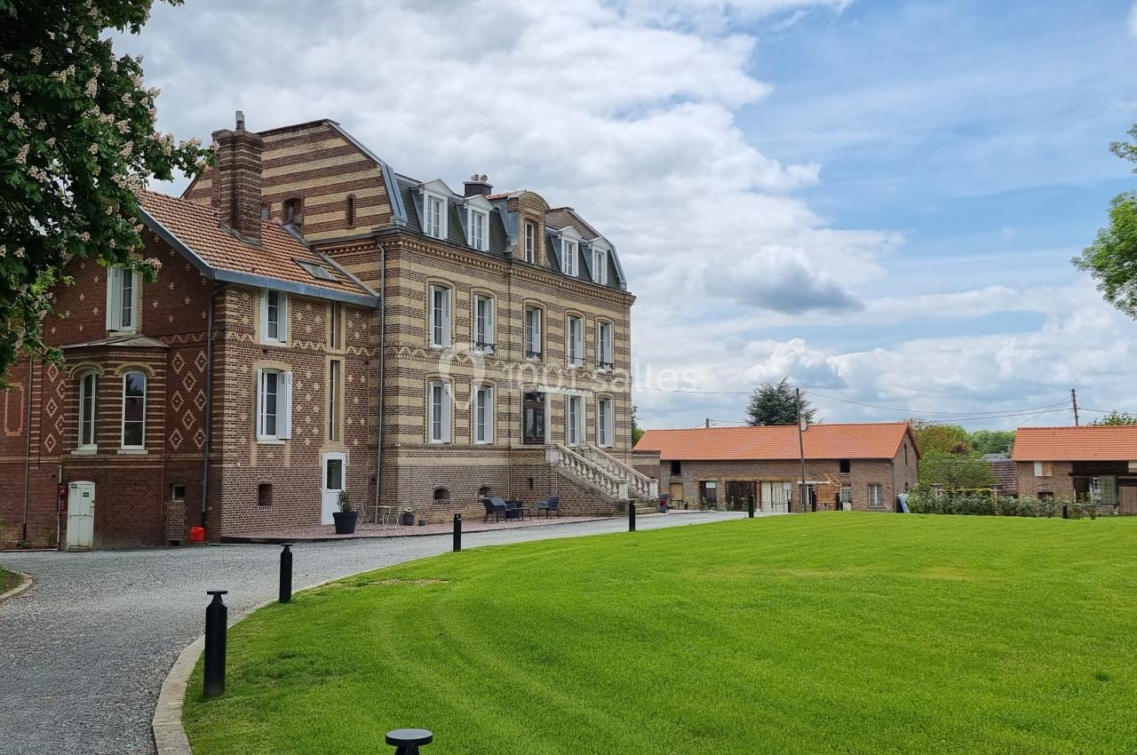 Bâtiment en briques rouges et beiges avec toit mansardé, entouré d'une pelouse et d'arbres sous un ciel partiellement…