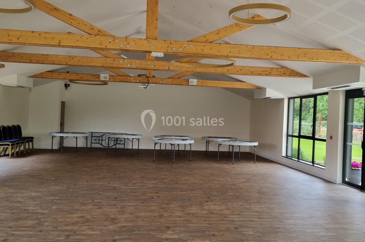 Salle lumineuse avec parquet, poutres apparentes, grandes baies vitrées et tables pliées contre les murs.