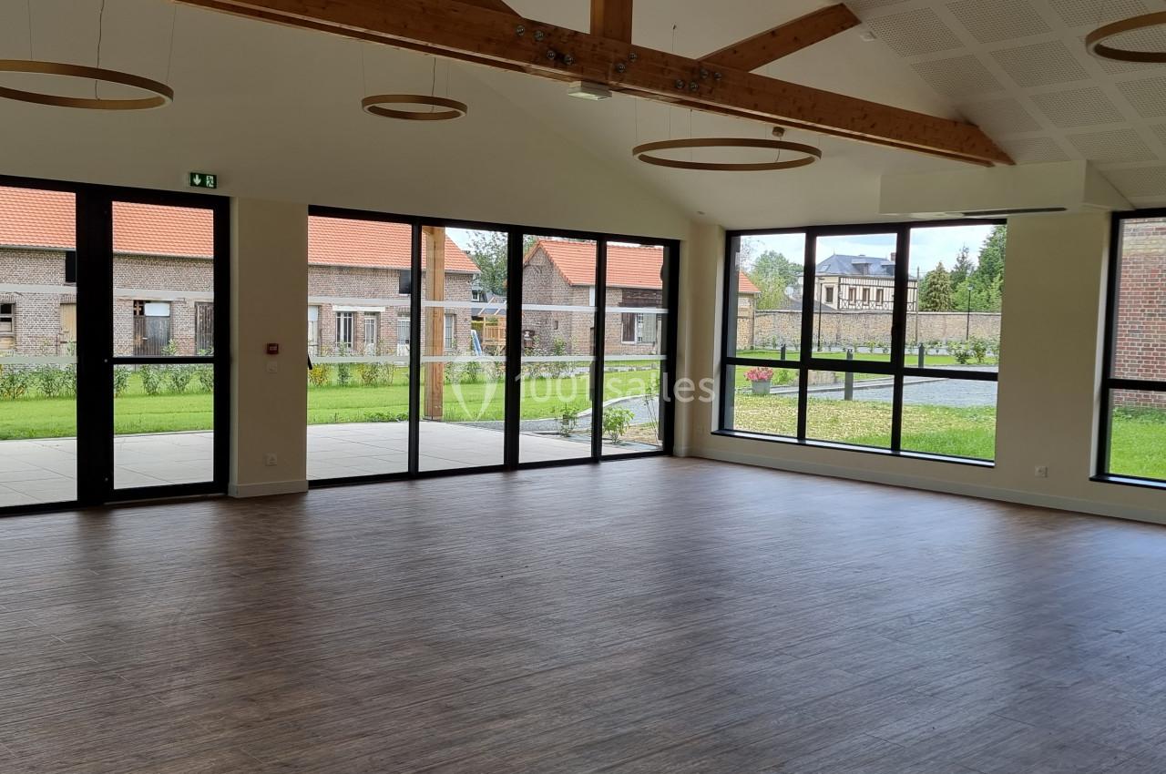 Salle lumineuse avec parquet, grandes baies vitrées donnant sur un jardin et poutres apparentes au plafond.