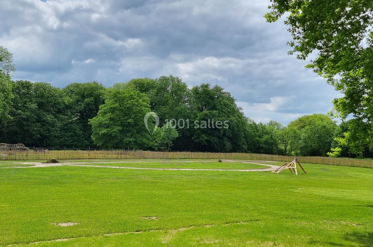 Grande étendue d'herbe entourée d'arbres, avec une structure en bois et un ciel partiellement nuageux.
