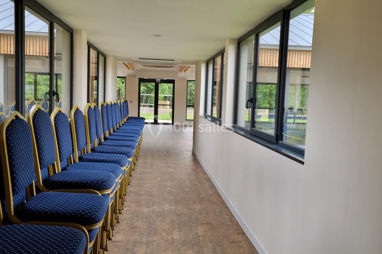 Rangée de chaises bleues alignées dans un couloir lumineux avec fenêtres donnant sur un espace extérieur verdoyant.