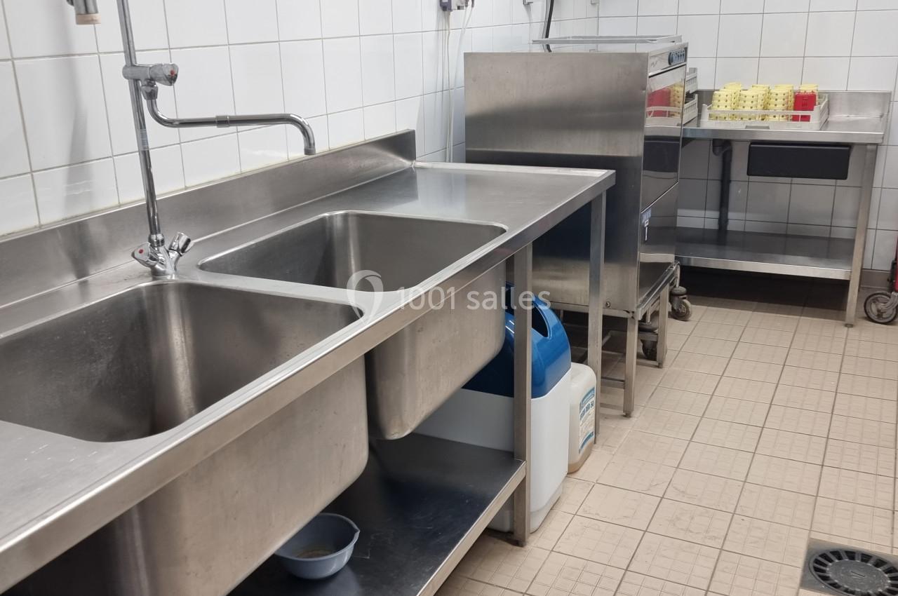 Évier double en inox dans une cuisine professionnelle avec étagères, chariot et équipements de nettoyage.