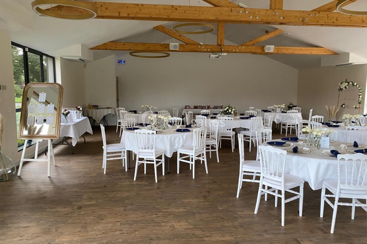 Salle de réception lumineuse avec tables rondes dressées, chaises blanches et décoration florale élégante.