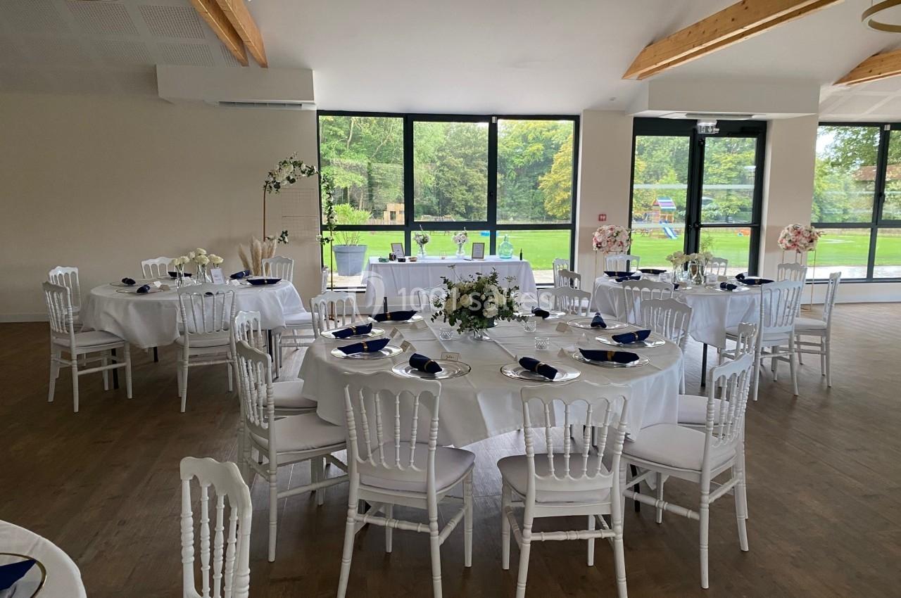 Salle de réception lumineuse avec tables rondes dressées, chaises blanches et vue sur un jardin verdoyant.