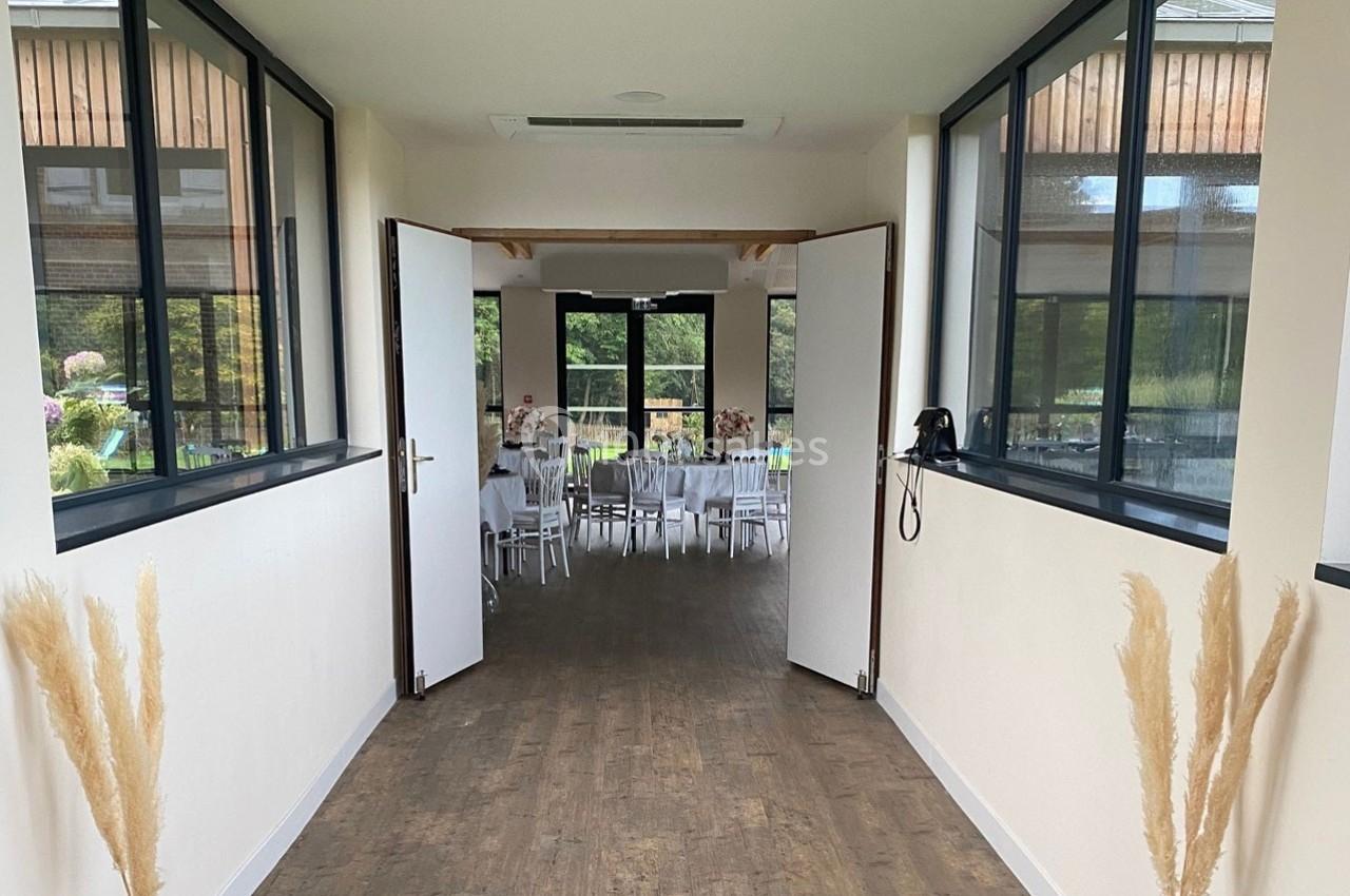 Vue d'un couloir lumineux menant à une salle de réception avec des tables dressées et des baies vitrées donnant sur un…
