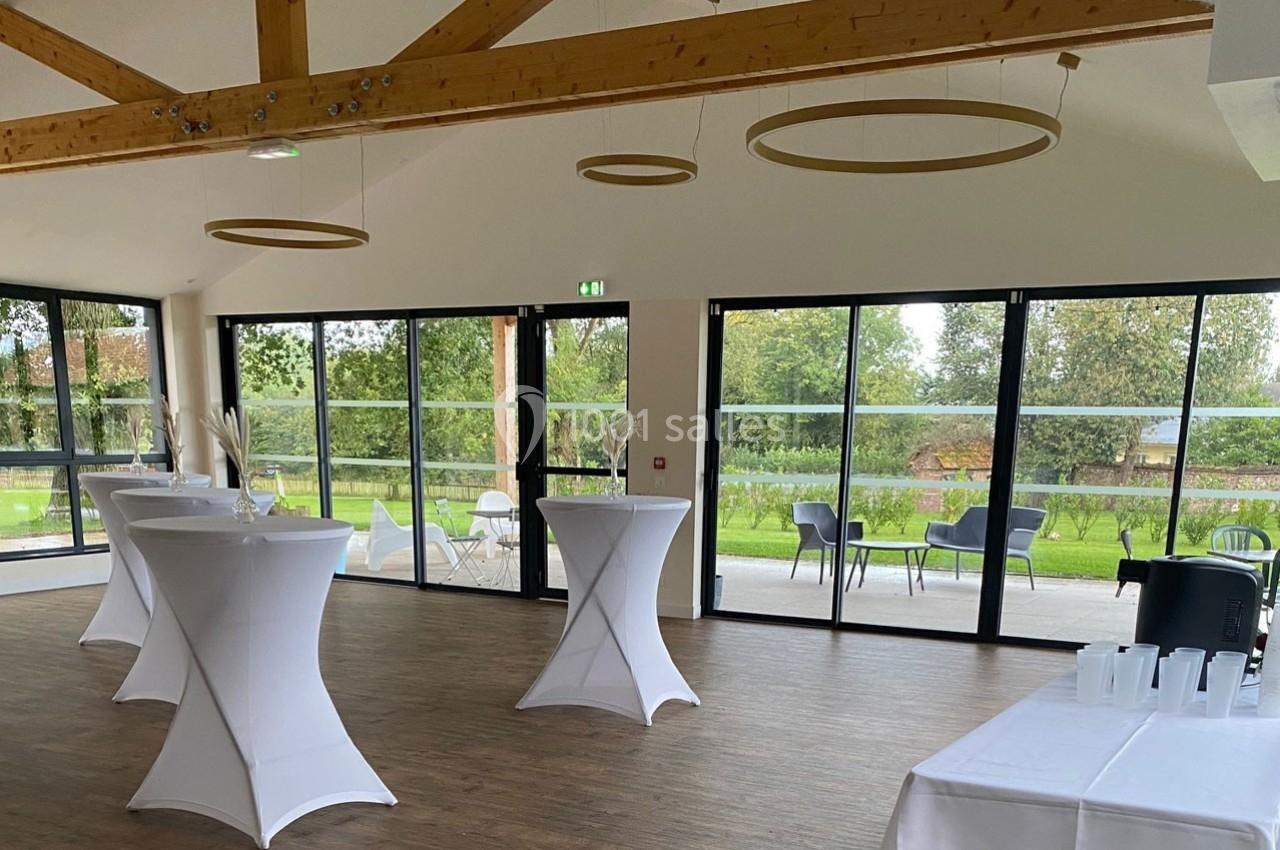 Salle lumineuse avec grandes baies vitrées, tables hautes nappées de blanc et vue sur un espace extérieur verdoyant.