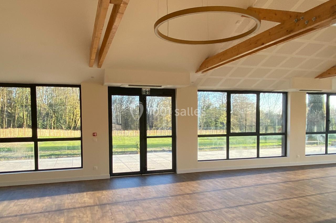 Grande salle lumineuse avec parquet, poutres apparentes et larges baies vitrées donnant sur un espace extérieur arboré.