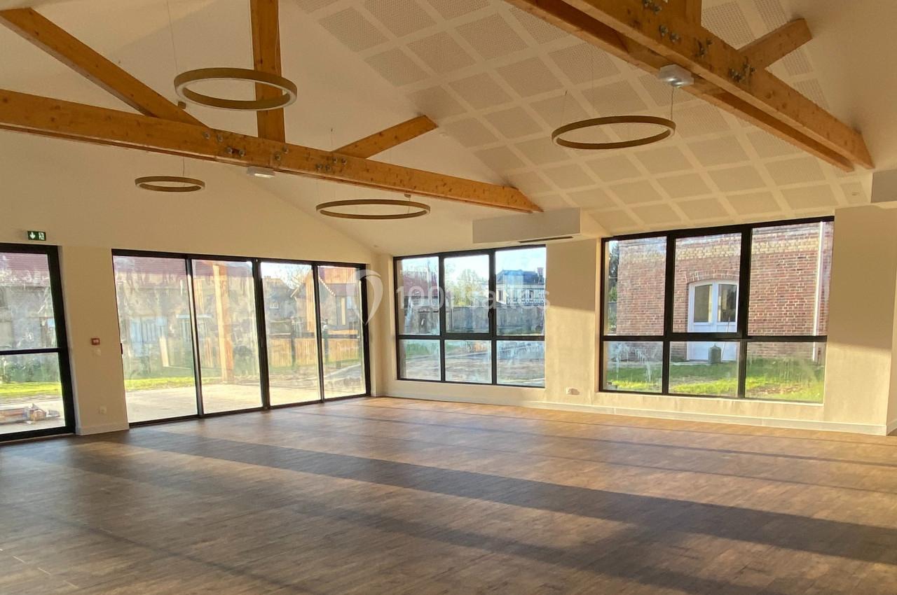 Salle lumineuse avec parquet, grandes baies vitrées, poutres apparentes et vue sur un extérieur verdoyant.