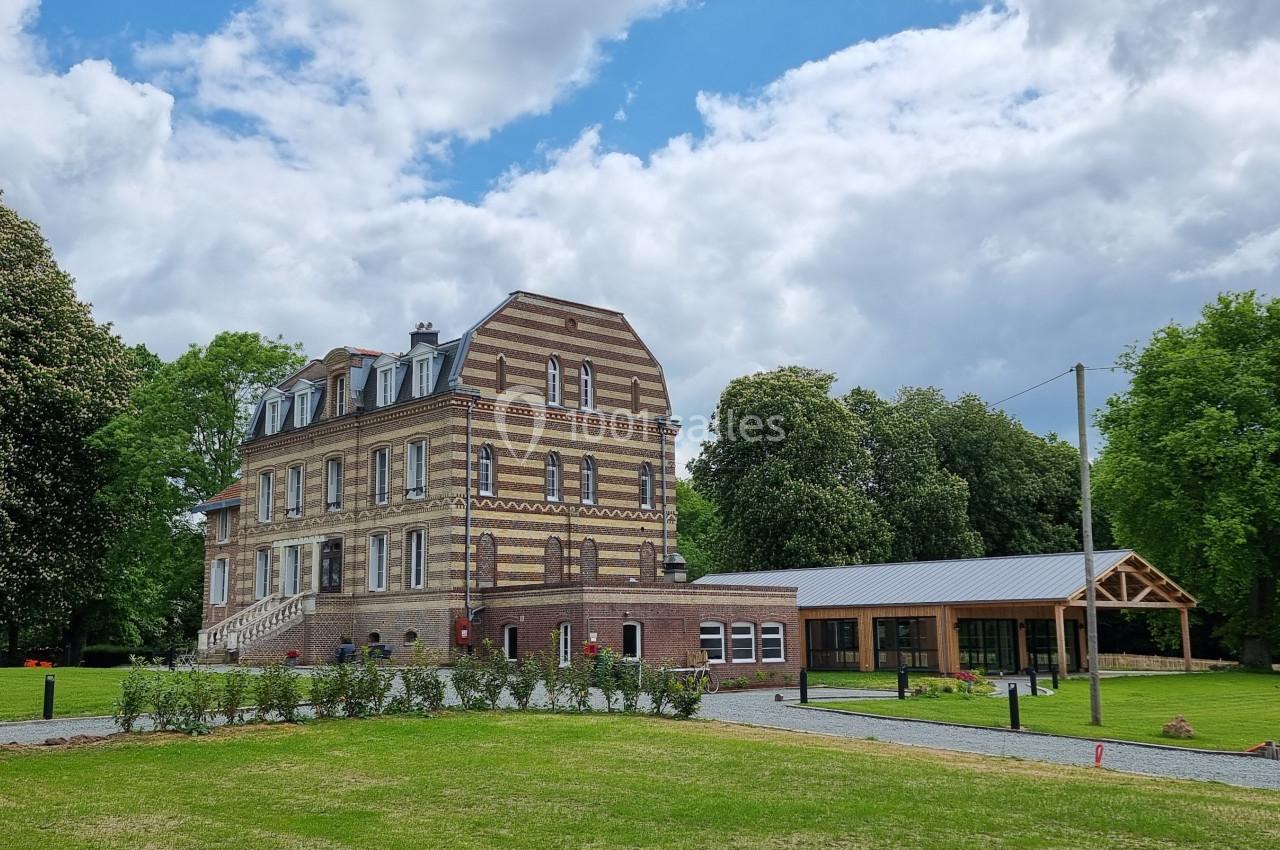 Manoir en briques entouré d'un jardin verdoyant, avec une annexe moderne en bois et un ciel partiellement nuageux.