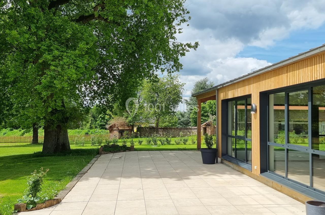 Terrasse en dalles claires bordant un bâtiment en bois avec de grandes baies vitrées, entourée d'arbres et de verdure.