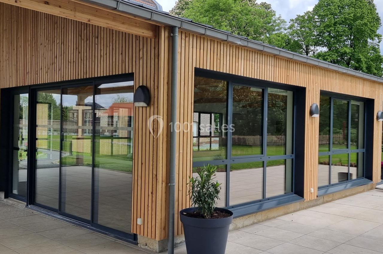 Bâtiment moderne avec façade en bois, grandes baies vitrées et terrasse dallée entourée de verdure.
