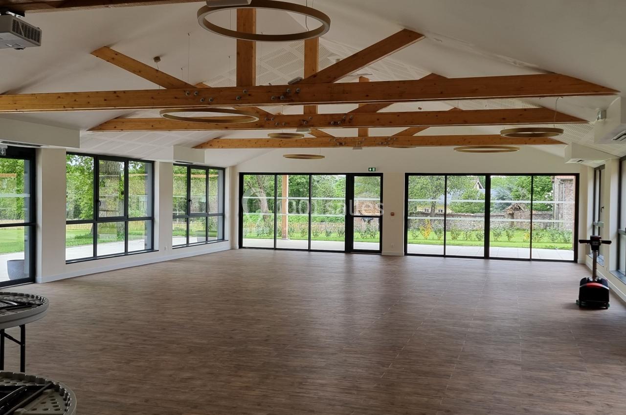 Salle lumineuse avec parquet, poutres apparentes, grandes baies vitrées donnant sur un espace extérieur verdoyant.