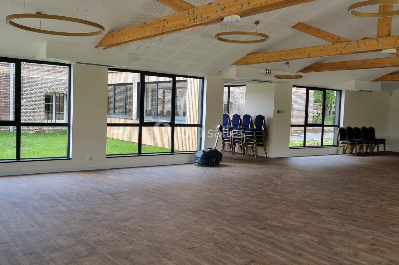 Salle lumineuse avec parquet, grandes fenêtres donnant sur un jardin, poutres apparentes et chaises empilées dans un coin.