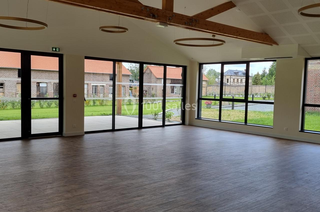 Salle lumineuse avec parquet, grandes baies vitrées donnant sur un jardin et poutres apparentes au plafond.