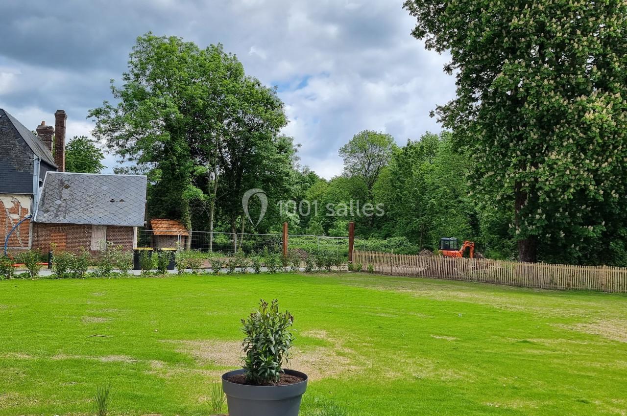 Jardin avec pelouse, arbres, clôture en bois et une maison ancienne en arrière-plan sous un ciel nuageux.