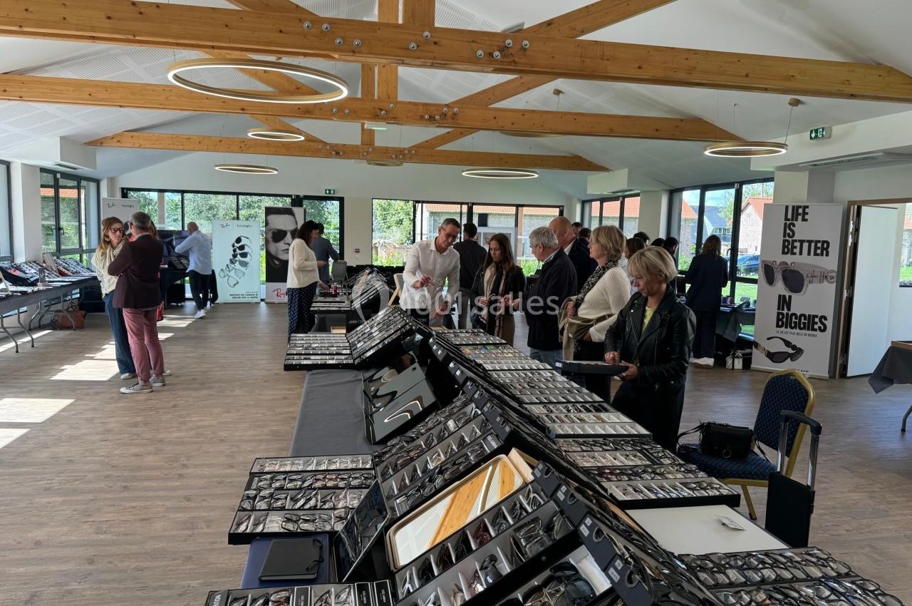 Des personnes examinent des montures de lunettes exposées sur des tables dans une salle lumineuse avec poutres en bois.